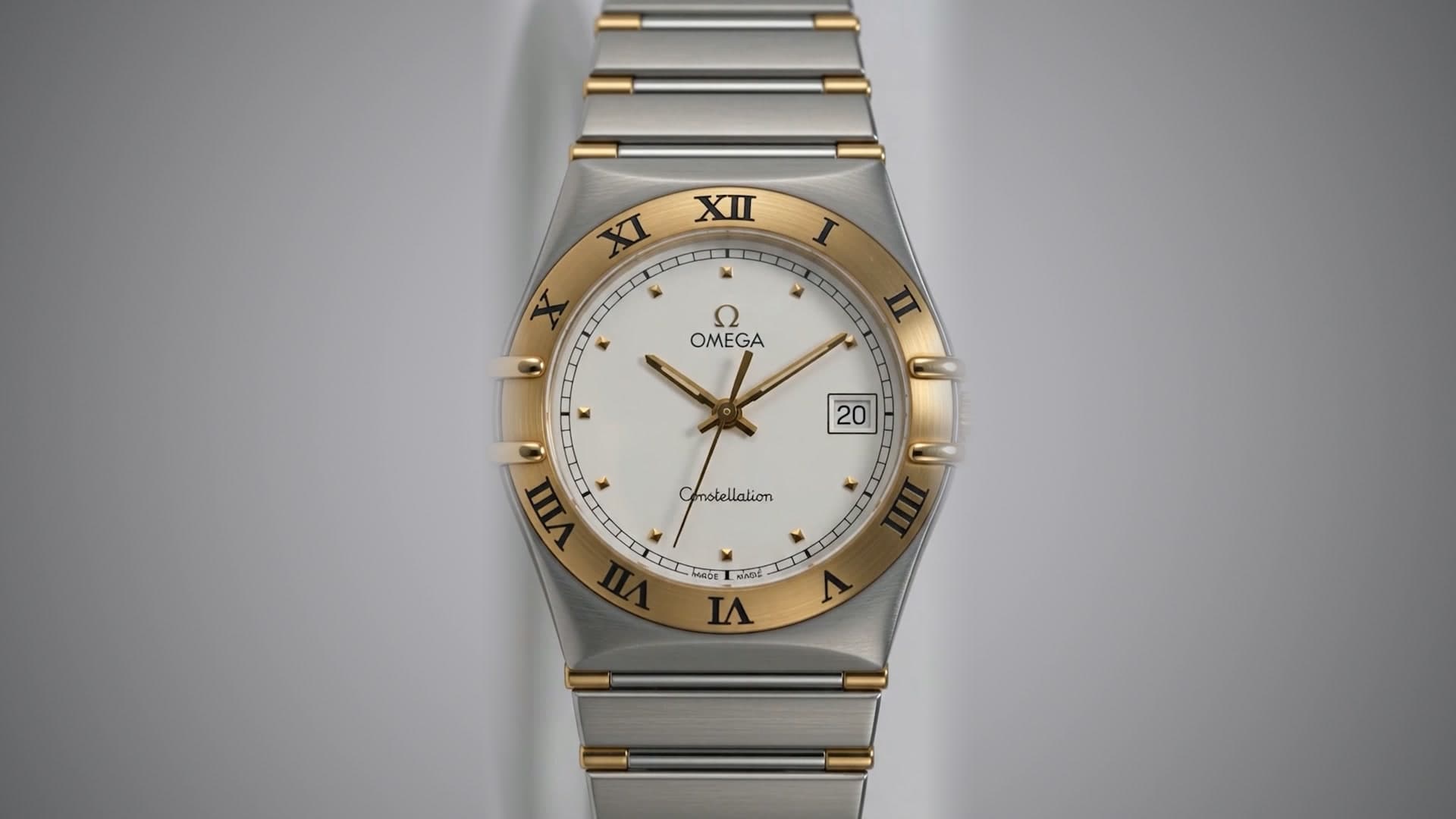 A two-tone watch with a white dial and a gold Roman-numeral bezel, on a grey studio backdrop